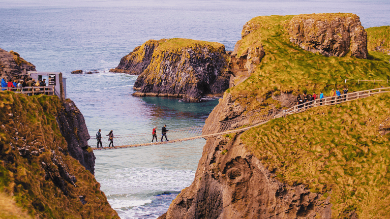 2 couples crossing Carrick-a-Rede Rope Bridge, with the crystal blue sea in the foreground. A small queue of visitors waiting to cross onto the island, and visitors queuing to get back other to the mainland.